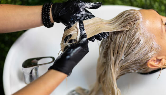 Hair Relaxer Lawsuit: A hairstylist wearing black gloves applying hair treatment or dye to a client's wet, blonde hair at a salon sink.
