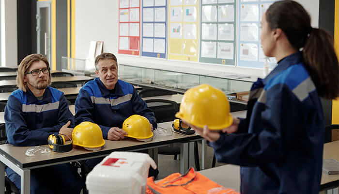 Industrial workers receiving training on safety regulations, wearing protective uniforms with hard hats and safety gear in a classroom setting.