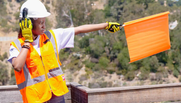Construction worker in high-visibility vest and hard hat following safety regulations while signaling with an orange flag on a job site.