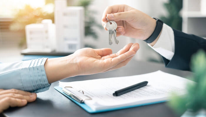 A person handing over keys to another individual, symbolizing the completion of a rental process, with a rental packet and documents on the table.