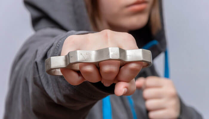 A person wearing a hoodie holding metal brass knuckles toward the camera, highlighting the question: Are brass knuckles legal in the U.S.?