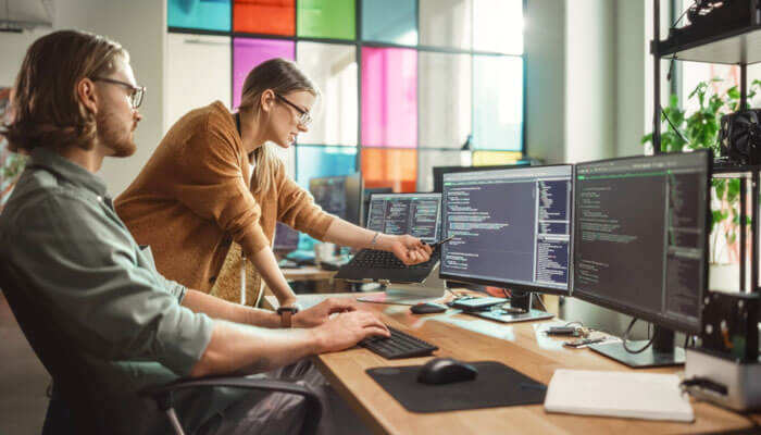 Two professionals collaborating at a desk with multiple monitors, working on business outsourcing tasks and coding solutions for clients.