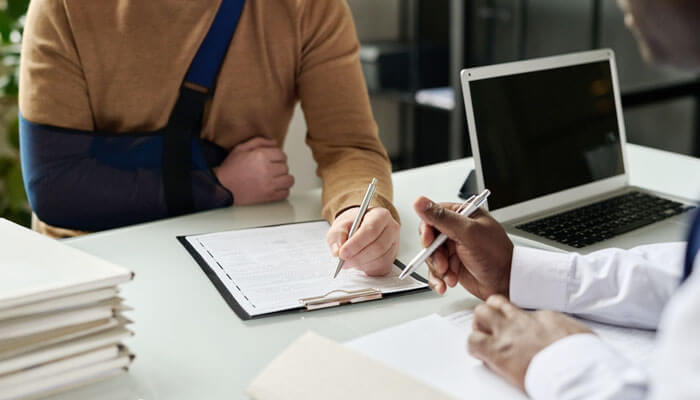 Injured person with arm in sling consulting a lawyer and signing documents related to Personal Injury Claims at a desk with files and a laptop.