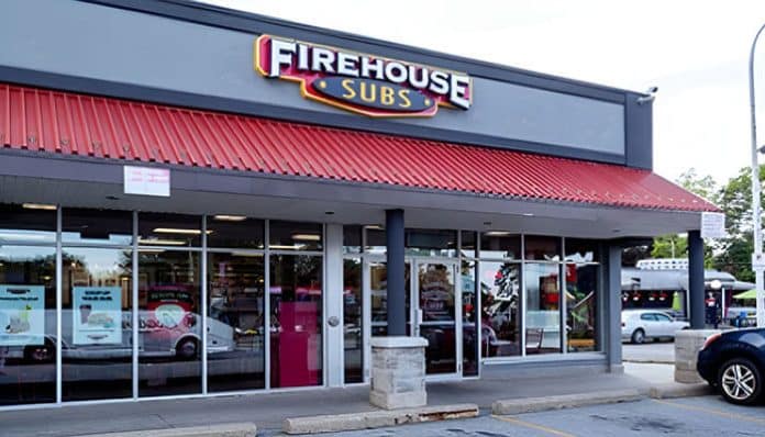 Exterior view of a Firehouse Subs restaurant with a red roof, large glass windows, and the Firehouse Subs logo sign displayed prominently above the entrance.