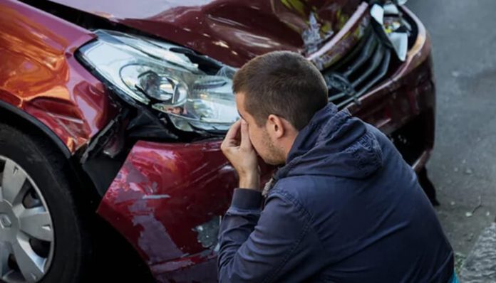 A person with short hair is kneeling beside a red car with a damaged front, holding his face in his hands, showing signs of distress following a car accident.