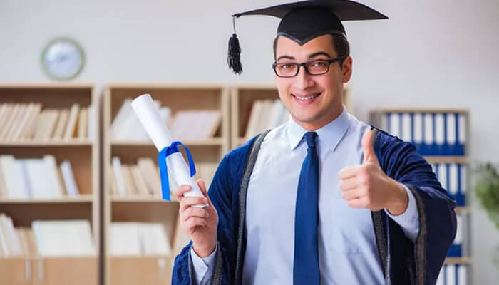 Smiling man in graduation gown and cap holding a diploma and showing thumbs up, representing the issue of fake degrees and their misleading appeal.