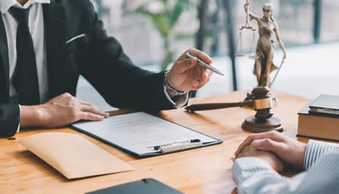 Law firm attorney discussing legal documents with a client at a desk, with gavel and Lady Justice statue in the background.