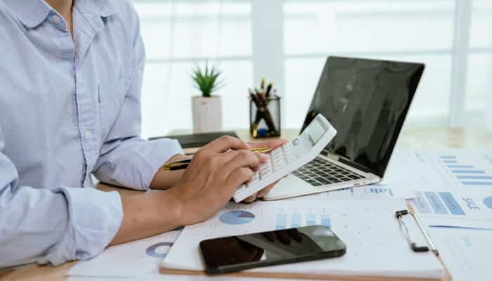 Person calculating budgets with a laptop, smartphone, and financial charts on a desk, illustrating future-proof planning and strategic decision-making.