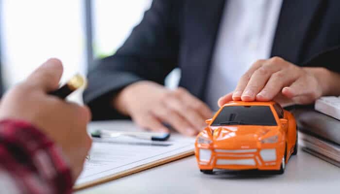 A person signing insurance documents while another places a hand on a small toy car, symbolizing claims assistance and legal support by motorcycle accident lawyers.