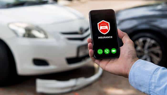 Person holding a smartphone with an insurance app open near damaged cars, symbolizing how to file and track a Car Accident Claim after a crash.