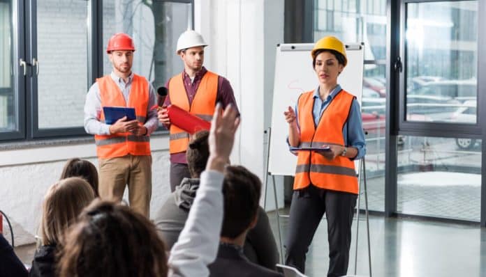 Workplace safety training session in progress with construction workers listening attentively to a female trainer explaining safety procedures and protocols.