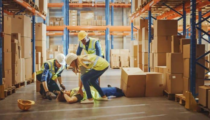 In a warehouse, three workers in hard hats and vests perform first aid on a person lying on the floor.