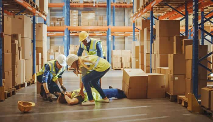 In a warehouse, three workers in hard hats and vests perform first aid on a person lying on the floor.