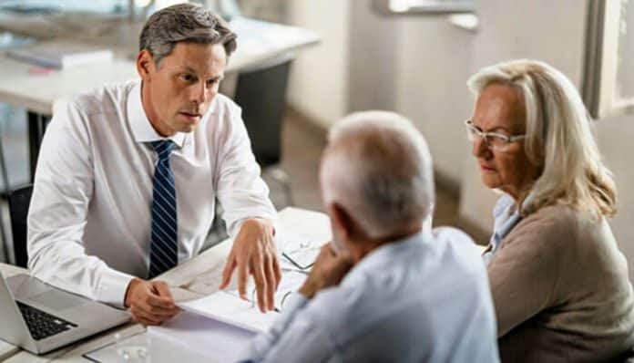 Man in a white shirt and blue tie discusses documents with two older adults at a table in an office environment.