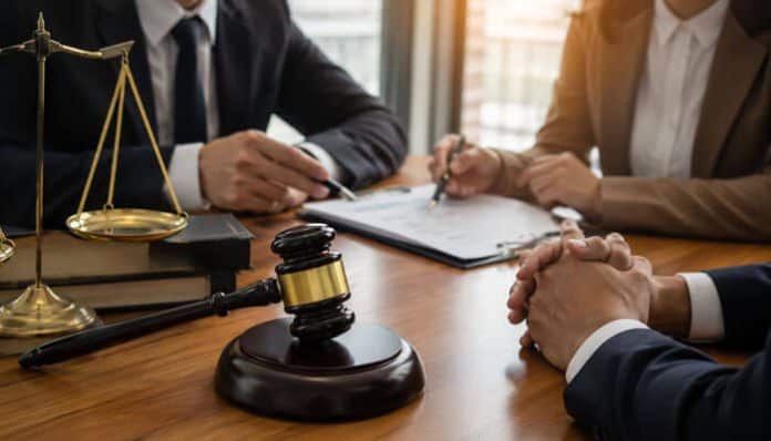 Law office scene with a gavel, scales of justice, and two professionals signing documents at a wooden table.