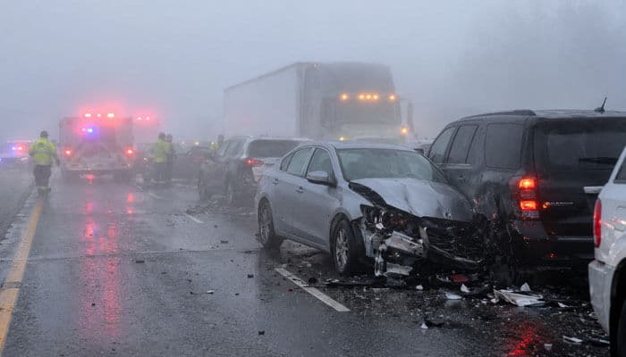 Common Accidents in Dense Fog - My Legal Opinion - My Legal Opinion White sedan with heavy front-end damage after a multi-vehicle crash on a rain-soaked highway; emergency responders and flashing emergency lights in foggy conditions behind a line of vehicles and a large truck.